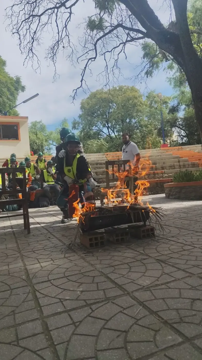 Voluntaria con casco verde, gafas y guantes avanza agachada con el extintor hacia el fuego real de práctica