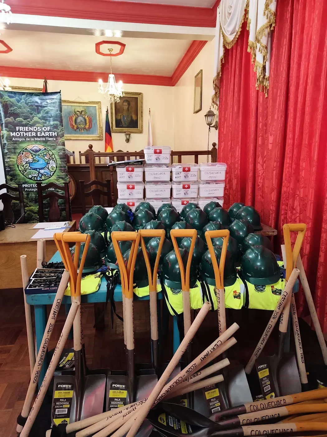 Mesa con cascos verdes, chalecos reflectivos, palas, silbatos y botiquines CERT junto al banner de Amigos de la Madre Tierra