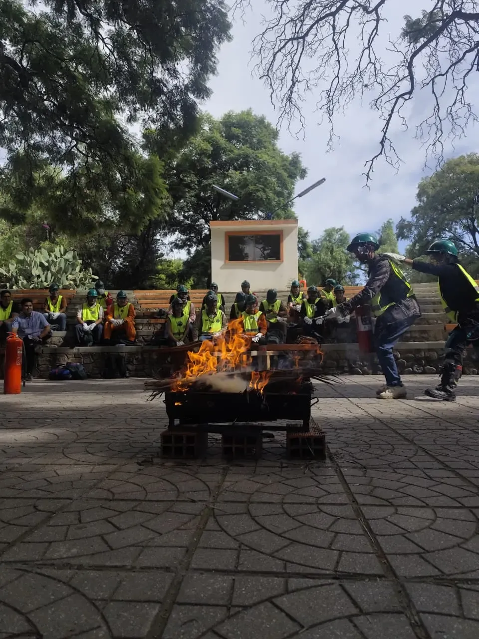 Llamas de la práctica de extintores con dos voluntarios equipados y el grupo observando desde las gradas