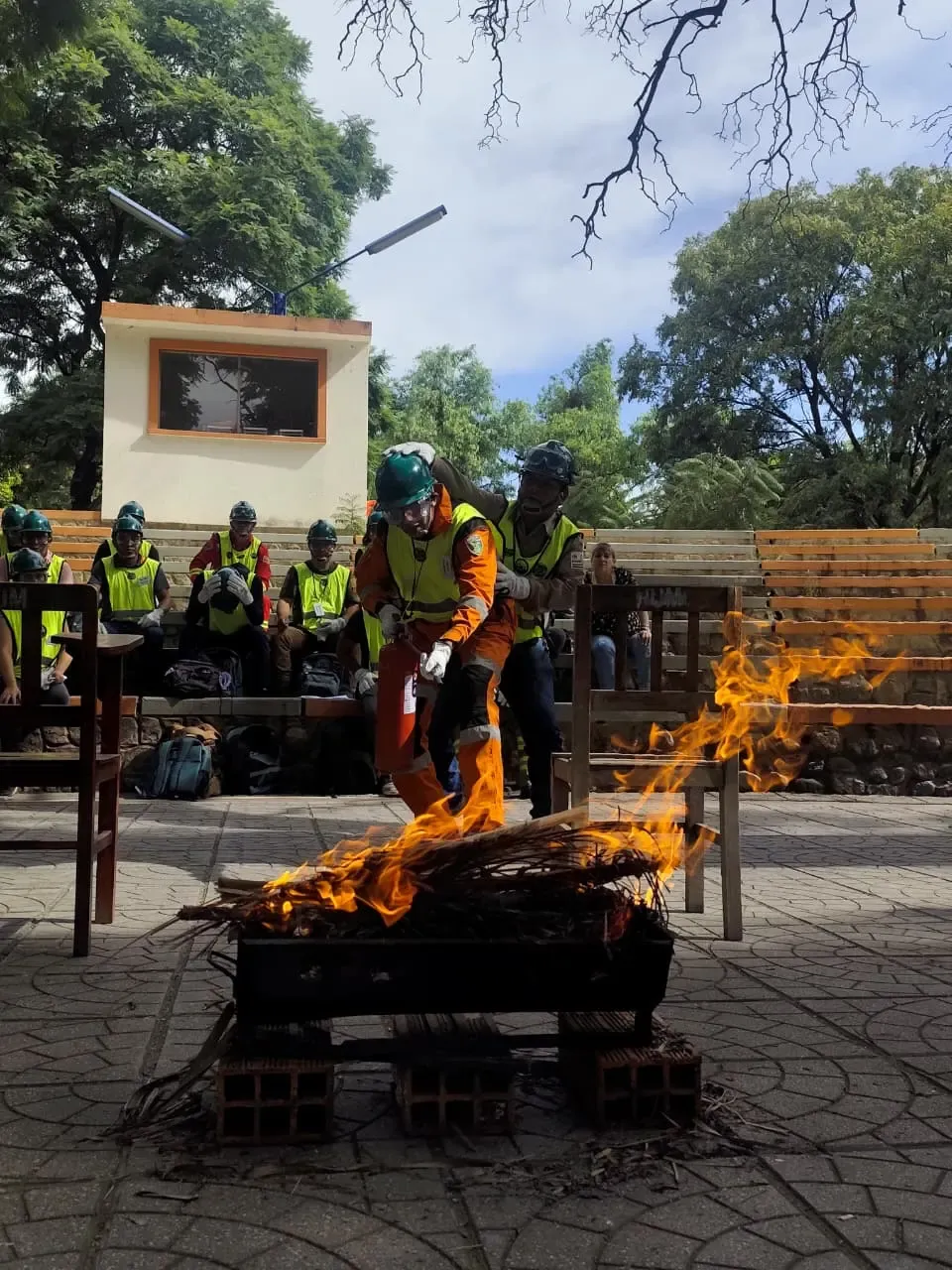 Dos brigadistas con cascos verdes y overol naranja avanzan con extintor hacia fuego real durante practica en anfiteatro, con companeros observando