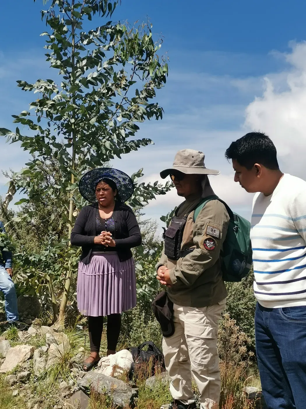 Mujer de pollera y sombrero comparte su testimonio ante un paramédico y un técnico junto a un eucalipto en la ladera