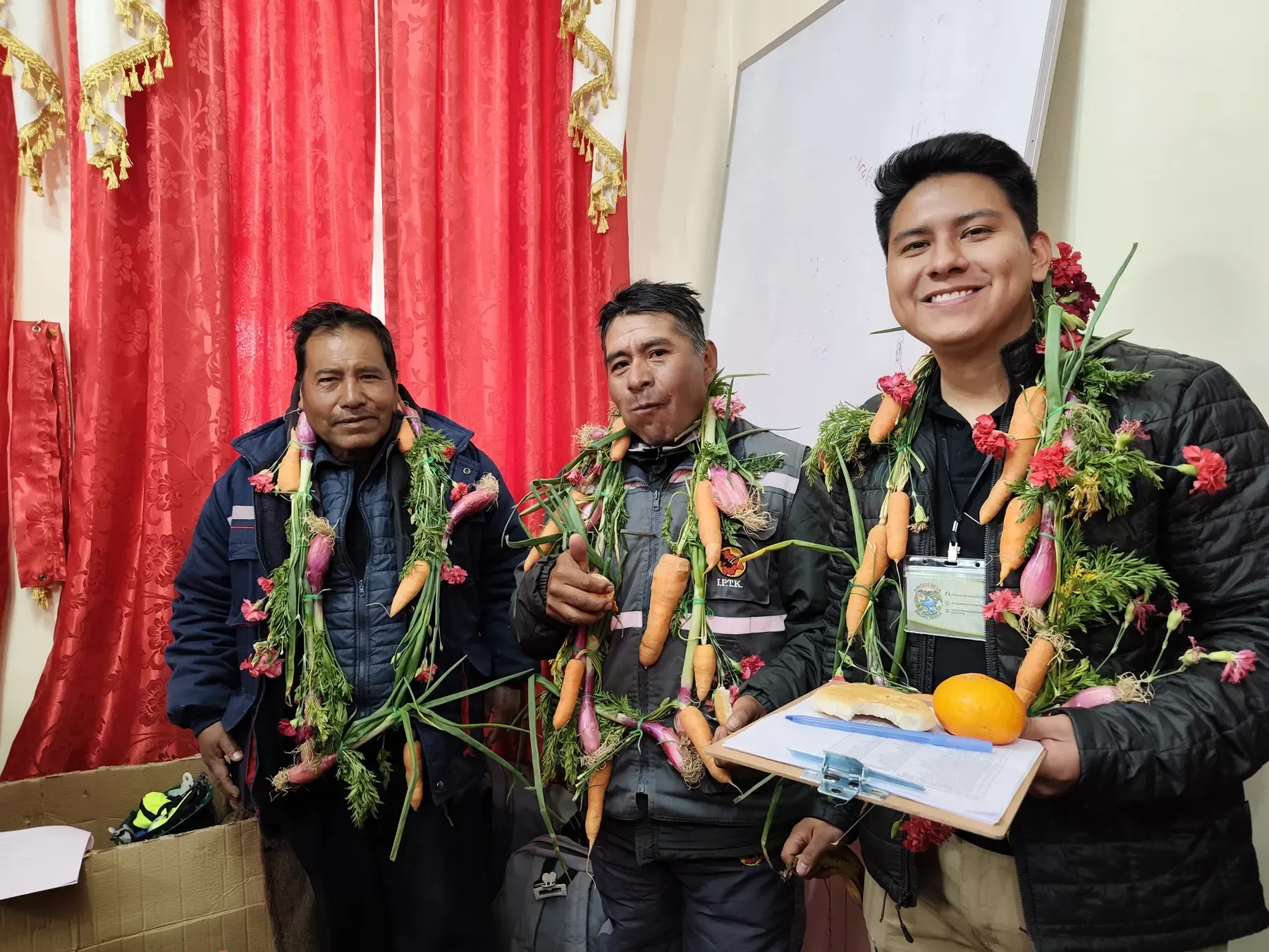 Tres facilitadores sonríen con guirnaldas de zanahorias, cebollas y claveles, el agradecimiento tradicional andino tras la capacitación CERT