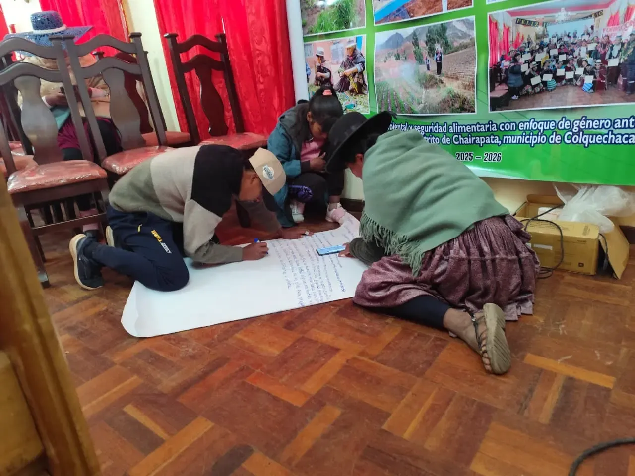 Participantes arrodillados escriben su plan comunitario en un papelógrafo bajo el banner del proyecto de resiliencia de Chairapata