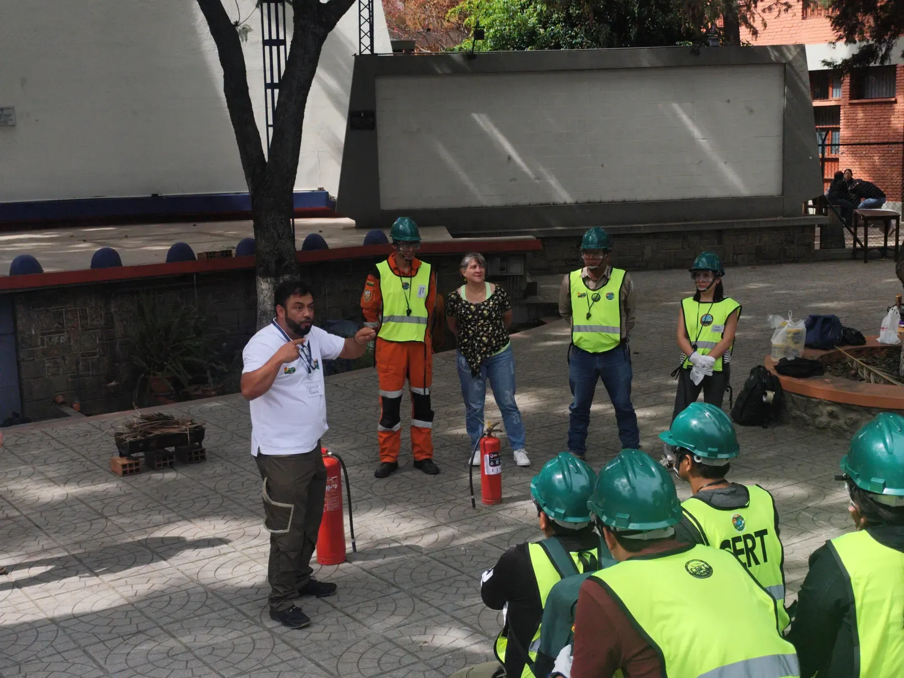 Instructor da indicaciones a voluntarios equipados junto a la parrilla preparada para la práctica de extintores