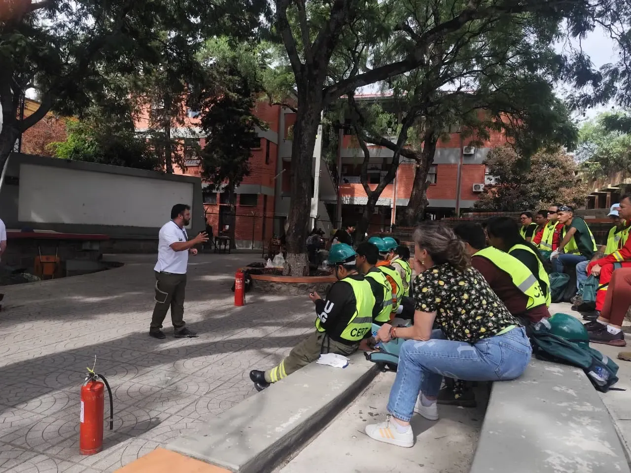 Instructor explica manejo de extintores a participantes con chalecos CERT sentados en gradas de un patio arbolado