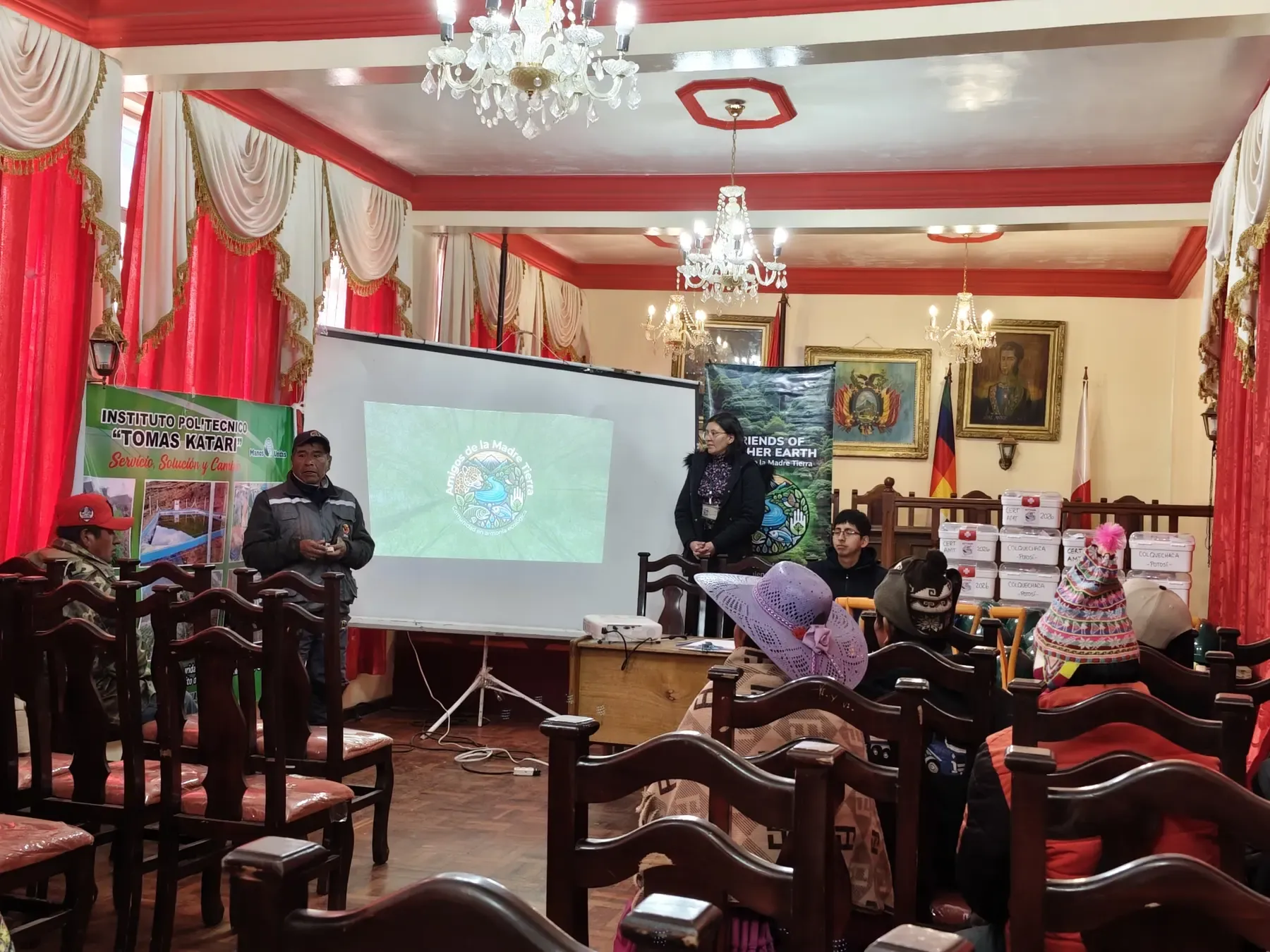 Acto de clausura en el salón municipal: autoridades y equipo de la ONG hablan ante comunarios con el logo de Amigos de la Madre Tierra proyectado