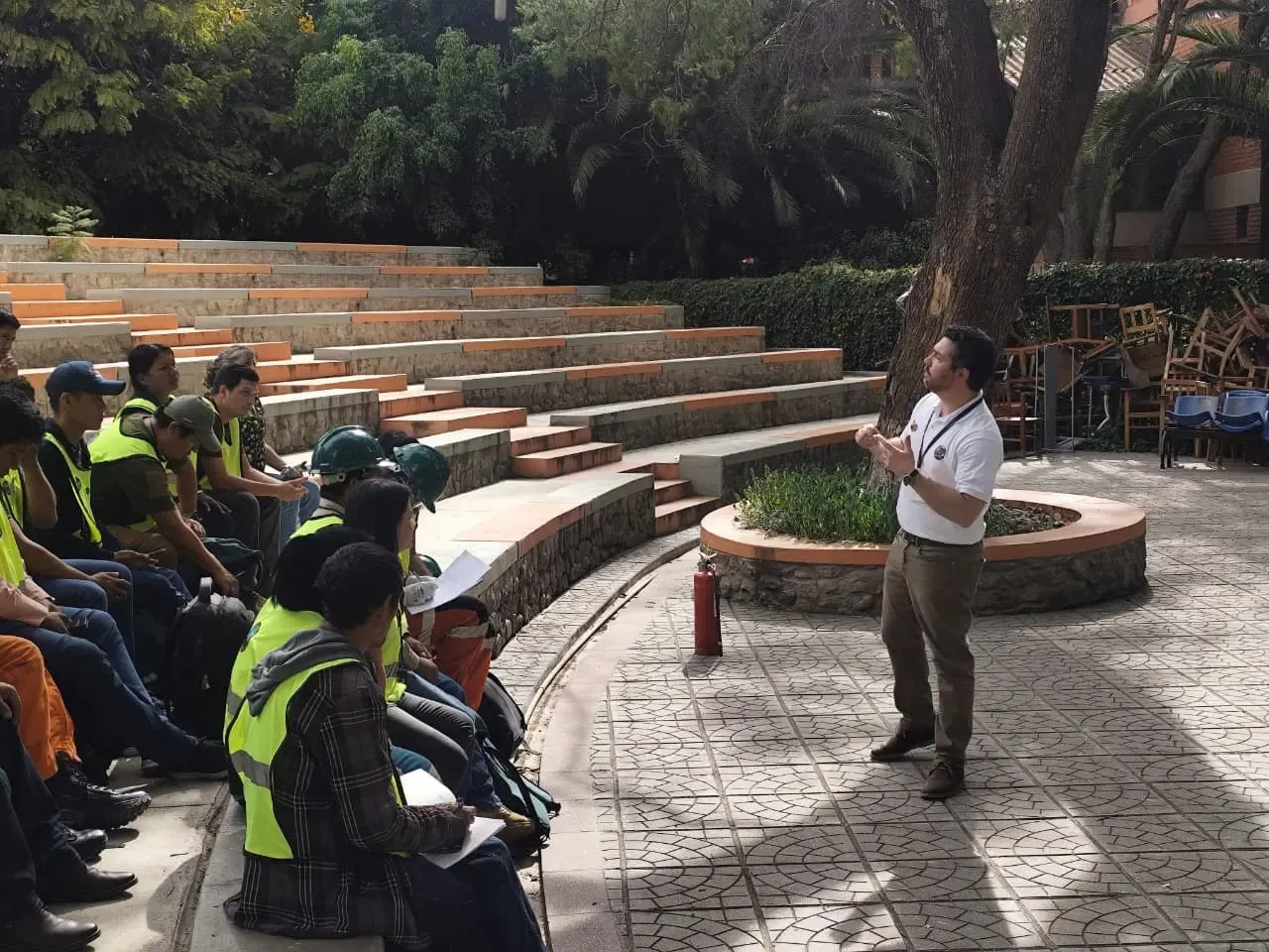 Vista amplia del anfiteatro: el instructor de pie explica ante el grupo sentado con cascos y chalecos, extintor en el centro