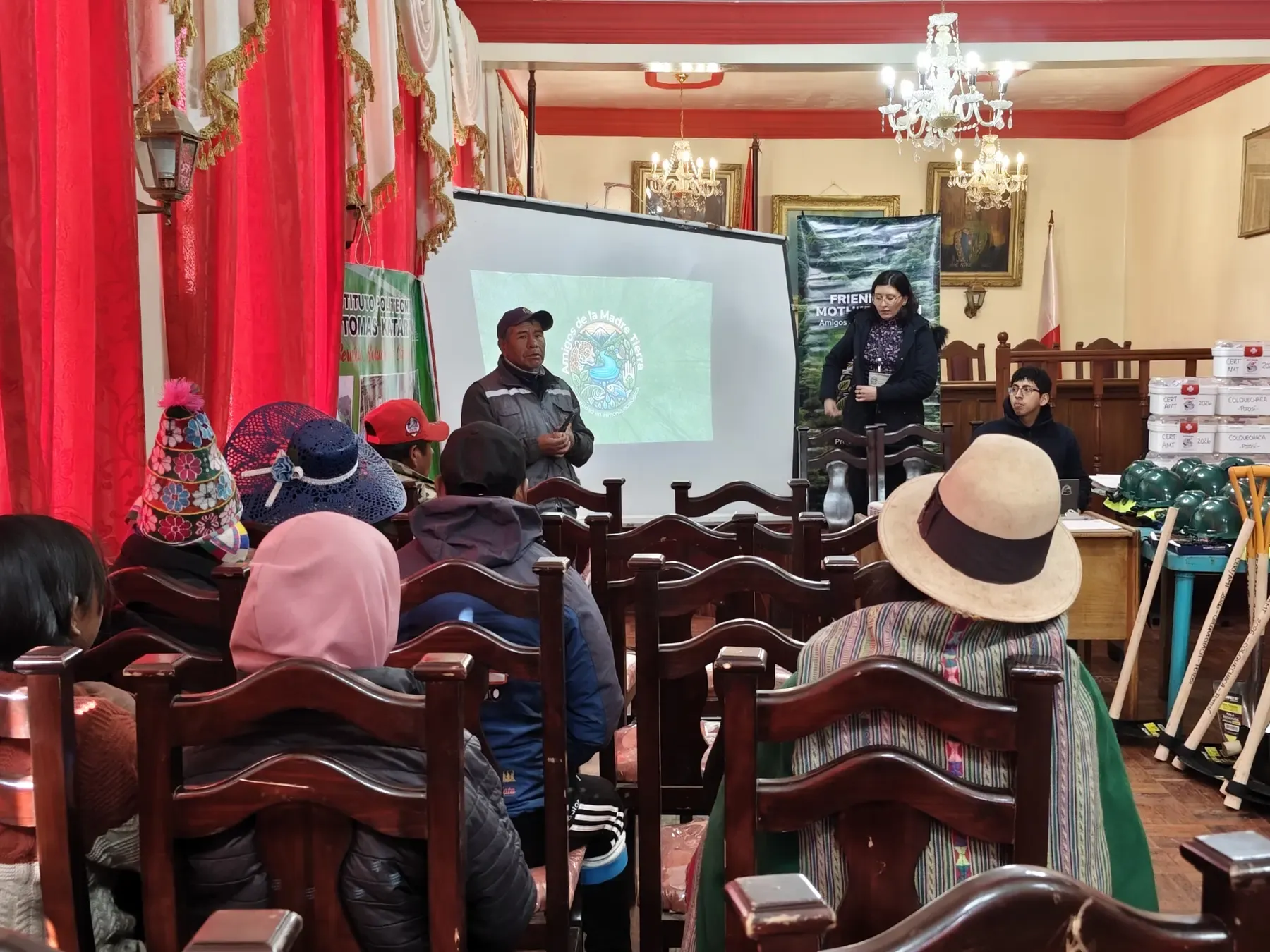 Dirigente comunal habla de pie ante el taller CERT con el logo de Amigos de la Madre Tierra proyectado y cascos verdes y botiquines listos a la derecha