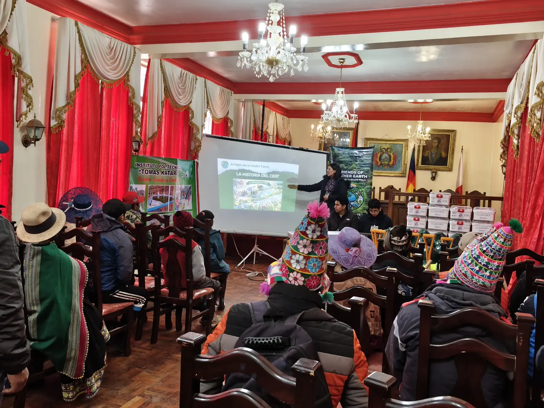 Facilitadora senala la diapositiva La Historia del CERT ante un salon lleno de comunarios con chullos y mantas andinas