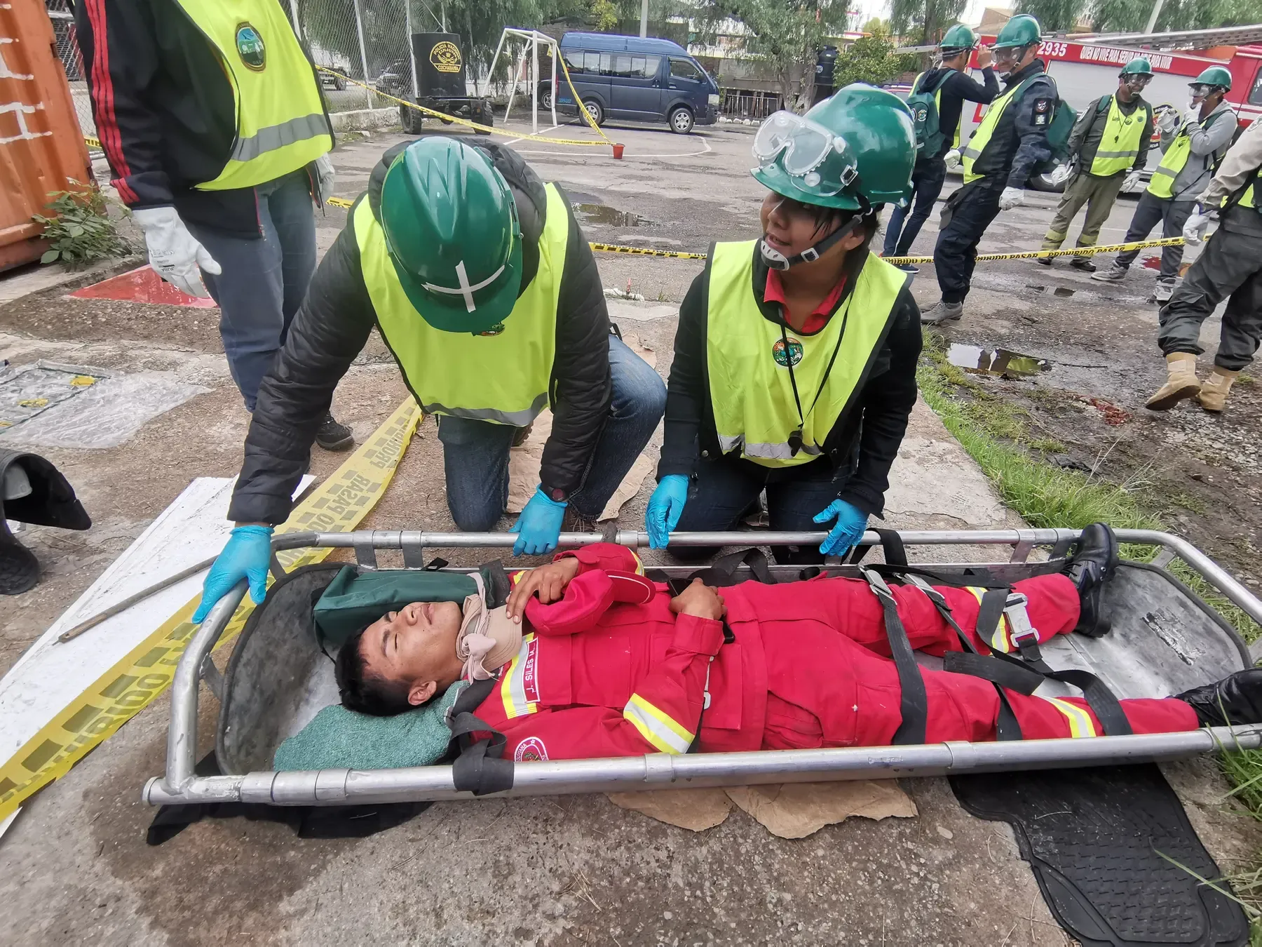 Dos voluntarios CERT con cascos verdes y guantes atienden a una víctima simulada inmovilizada en camilla durante el simulacro, con bomberos y cinta de seguridad al fondo